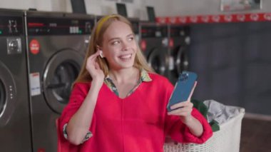 Çekici, güler yüzlü, genç, sarışın bir kadın telefon ve kulaklık kullanıyor. Çamaşır odasında oturmuş çamaşır yıkıyor. Ev işi çamaşır makinesi kurutma makinesi içeride. Kapatın. Yavaş çekim