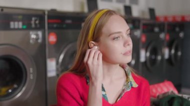Çamaşır makinesinin yanında oturan güzel sarışın kadın kameraya bakıyor ve kulaklıkla müzik dinliyor. Çamaşır odası ev işi. Yavaş çekim