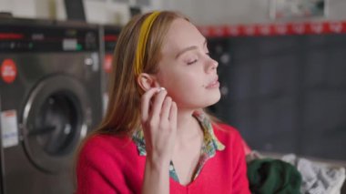 Gülümseyen genç sarışın kadın çamaşır makinesinin yanında oturuyor kameraya bakıyor ve kulaklıkla müzik dinliyor. Çamaşır odası ev işi. Yavaş çekim