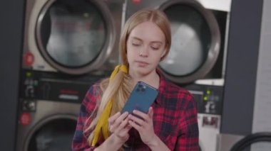 Genç, sarışın, gülümseyen bir kadın çamaşır odasında kareli gömlekle çamaşır yıkarken telefon kulübesini kullanıyor. Ev işi çamaşır makinesi kurutma makinesi içeride. Yavaş çekim