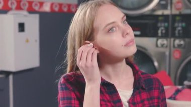 Gülümseyen yüzlü güzel sarışın kadın çamaşır makinesinin yanında oturuyor ve kulaklıkla müzik dinliyor. Çamaşır odası ev işi. Yavaş çekim