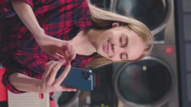 Dikey çekimde güzel sarışın kadın çamaşır makinesinin yanında oturuyor telefon kullanıyor ve kulaklıkla müzik dinliyor. Çamaşır odası ev işi. Yavaş çekim