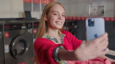 Çamaşır odasında fotoğraf çekerken gülümseyen ve akıllı telefon kamerasına poz veren güzel bir kadın. Ev işi çamaşır makinesi kurutma makinesi içeride. Kapatın. Yavaş çekim