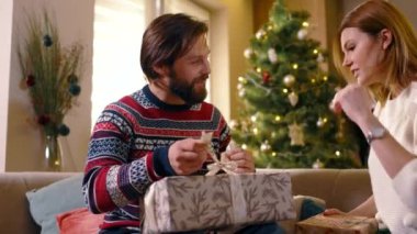 Mutlu sakallı adam yılbaşı ağacının yanındaki kız arkadaşının hediyesini açıyor. Heyecanlı evli Kafkasyalı çift tatillerini evde geçirerek hediye alışverişinde bulunuyor..