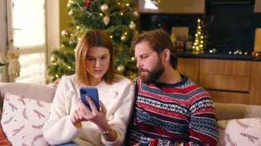 Noel tatili. Oturma odasında oturan beyaz erkek ve kız arkadaş çifti cep telefonu uygulaması kullanarak aile için hediyeler seçiyor..