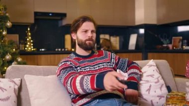 Sinirli, stresli, sakallı, beyaz bir adam oturma odasında televizyon seyrediyor, kanallara bakıyor ve sıkılıyor. Oturma odası. Daire. Kış tatili.