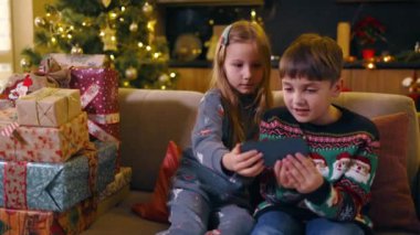 Beyaz tenli iki çocuk oturma odasında oturmuş akıllı telefon internet bağlantısı kullanıyor. Noel arifesinde internetten komik uygulamalar oynuyor..