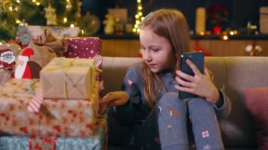 Mutlu Kafkasyalı küçük kız video sohbetleri akıllı telefon kayıtlarında hediyelerle dolu kutuları gösteriyor evde noel ağacının yanında eğleniyor..