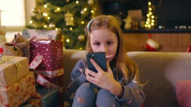 Küçük sevimli kız, Noel 'de evde kalıp sınıf arkadaşlarıyla cep telefonunda sohbet ederek bir sürü hediye gösteriyor. Tatillerde. Çocukluk.