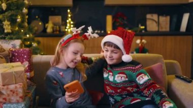 İki güzel genç kız ve erkek kardeş tatil şapkası takıp gülümsüyor ve akıllı telefondan selfie çekiyor. Noel ağacı. Noel ailesi.