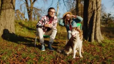 Gülümseyen mutlu çiftimiz güz parkında ya da ormanda evcil bir köpekle oturuyor. Hayvan gibi rahat, neşeli bir portre. Gün ışığında. Yavaş çekim