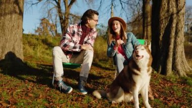 Güz parkında ya da ormanda evcil bir köpekle oturan çekici mutlu çift. Hayvan gibi rahat, neşeli bir portre. Gün ışığında. Yavaş çekim