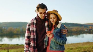 Güzel genç bir çift, göl kenarındaki ormanda telefon kullanıyor. Mutlu bir şekilde gülümsüyorlar. Dışarıdaki sosyal ağlar. Portre. Kapatın. Yavaş çekim
