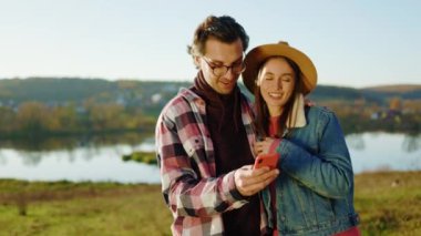 Şirin genç bir çift göl kenarındaki ormanda telefon kullanıyor. Gülümsemekten mutlu oluyorlar. Dışarıdaki sosyal ağlar. Portre. Kapatın. Yavaş çekim