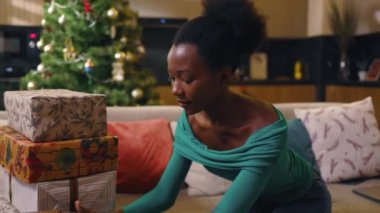 Gülümseyen mutlu Afro-Amerikan güzel genç bayan koltukta oturuyor ve dekore edilmiş odada Noel hediyesi kutusunu hazırlıyor. Yeni yıl. Tatil kutlaması. Noel ağacı. Hediye. Yavaş çekim