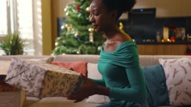 Mutlu Afro-Amerikan güzel genç bayan koltukta oturuyor ve dekore edilmiş odada Noel hediyesi kutusunu hazırlıyor. Yeni yıl. Tatil kutlaması. Noel ağacı. Hediye. Yavaş çekim