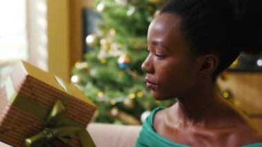 Kanepede oturan Afro-Amerikalı güzel genç bir kadın Noel hediyesi kutusunda dekore edilmiş odadaki kamera gülüşüne bakın. Yeni yıl. Tatil kutlaması. Noel ağacı. Hediye. Yavaş çekim