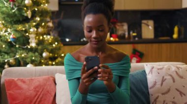Yakın plan Afro-Amerikan güzel genç bayan kanepede oturuyor ve akıllı telefonda daktilo kullanıyor. Mutlu ol. Yeni yıl gecesi. Tatil kutlaması. Noel ağacı. Hediye. Yavaş çekim
