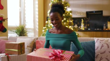 Afro-amerikan güzel, mutlu, genç bir kadın kanepeye oturmuş, gülücüklü konuşmalara bakıyor, Noel hediyelerini süslü odada tutuyor. Yeni yıl. Tatil kutlaması. Noel ağacı. Blog. Yavaş