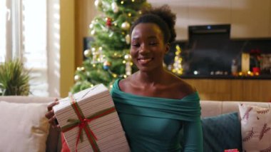Çekici Afrikalı Amerikalı güzel, mutlu, genç bir kadın kanepeye oturmuş, Noel hediyesini süslü odada tutarken gülümseyen bir gülümsemeye bakıyor. Yeni yıl. Tatil kutlaması. Noel ağacı. Yavaş çekim