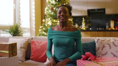 Afrika kökenli Amerikalı güzel bir genç kadın kanepeye oturmuş elini sallayıp gülümseyen konuşmasına bakıyor süslü bir odada Noel hediyesi kutusunu tutuyor. Yeni yıl. Tatil kutlaması. Blog. Yavaş çekim