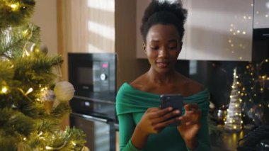 Mutlu Afrikalı Amerikalı genç bir kadının portresi Noel ağacının yanında duruyor ve cep telefonuna mesajlar yazıyor. Noel zamanı. Kış büyülü atmosferi kutluyor. Yavaş çekim