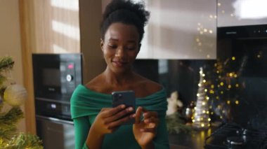 Mutlu Afrikalı Amerikalı genç kadın Noel ağacının yanında duruyor ve cep telefonuna mesajlar yazıyor. Noel zamanı. Kış büyülü atmosferi kutluyor. Yavaş çekim