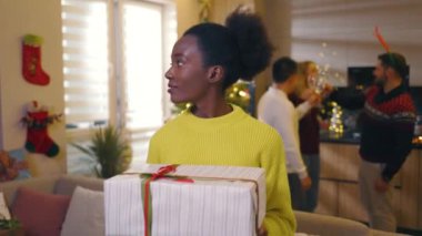 Afro-Amerikan güzel, mutlu, genç bir kadın. Gülümsemesine bakın. Noel hediye kutusunu dekore edilmiş odada tutun. Yeni yıl. Tatil kutlaması. Noel ağacı. Yavaş çekim