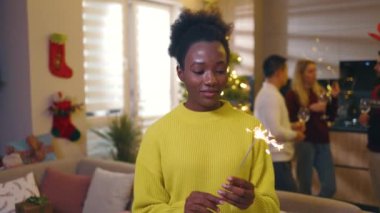 Noel ağacında süslü bir odada elinde maytapla gülümseyen geleneksel bayrama bakan Afrikalı Amerikalı genç bir kadın. Kutlama. Mutlu noeller. Kış zamanı. Yavaş çekim