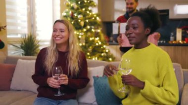 Çok ırklı iki kadın arkadaş noel partisi yapıyorlar ellerinde şarap tutuyorlar ev dansında kanepede oturuyorlar. Geleneksel tatil. Kutlama. Mutlu noeller. Kış zamanı. Yavaş çekim
