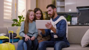 Kanepede bavulla oturan mutlu bir aile tablet bilgisayar kullanarak gülüyor ve evde birlikte eğleniyorlar. İnternet turizmi modern online seyahat. Kapatın. Yavaş çekim