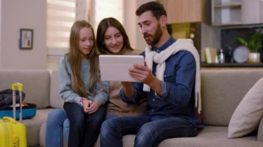 Çekici, genç bir aile bavullarıyla kanepede oturup tablet bilgisayar kullanarak gülüyor ve evde birlikte eğleniyorlar. İnternet turizmi modern online seyahat. Kapatın. Yavaş çekim