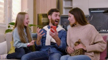 Aile telefonlarına bakıyor gülümseyerek gülüyor sosyal medya uygulamasında kazanıyor evde kanepede oturuyor. Gadget aşk eğlencesi akıllı telefon kutlaması. Yavaş çekim
