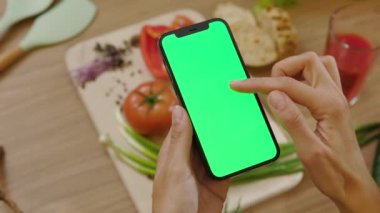 Kadın elleri ahşap tablet arka plandaki sebzelerin üzerinde dikey yeşil ekranlı telefonu tutuyor. Sağlıklı yemek mutfağı. Vejetaryen blog yazarı. Yavaş çekim