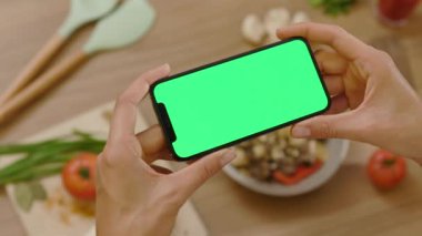 Genç elli kadın yatay yeşil ekranlı dokunmatik telefon kullanıyor. Ahşap tabletteki arka plan sebzeleri. Sağlıklı yemek mutfağı. Vejetaryen blog yazarı. Yavaş çekim