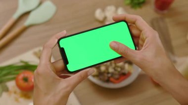 Ağır çekimde çalışan kadın yatay yeşil ekranlı dokunmatik telefon kullanıyor. Ahşap tabletteki arka plan sebzeleri. Sağlıklı yemek mutfağı. Vejetaryen blog yazarı..