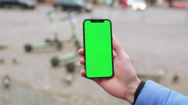 Elinde yeşil ekran akıllı telefonu olan genç bir adam arka plandaki elektrikli scooterlara dokunuyor ve onları yürütüyor. İnternet iletisi maketi görüntüsü. Yavaş çekim