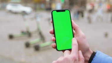 Yeşil ekran akıllı telefonlu bir adamın sokak arkaplanındaki elektrikli scooterlara dokunup sürtündüğü bir fotoğraf. İnternet iletisi maketi görüntüsü. Yavaş çekim