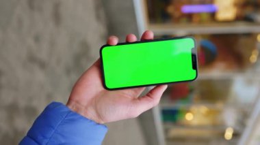 Elinde yeşil ekran akıllı telefonlu genç adam sokak arka planında bir yere dokunup duruyor. İnternet iletisi maketi görüntüsü. Dikey çekim. Yavaş çekim