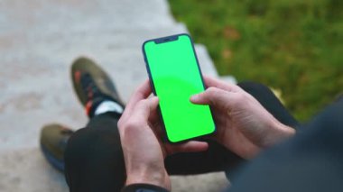 Erkek elinin omzunun üstünden yaklaş. Açık havada dikey yeşil ekranlı telefon kullan. Oturuyorum. Dokunmatik ekranlı internet cep telefonu kaydırması. Yavaş çekim