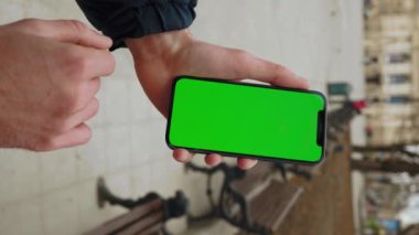 Dikey çekimde, arka plandaki bankta dikey yeşil ekranlı telefonu tutan adam görülüyor. İnternet parmak aleti. Kapatın. Yavaş çekim