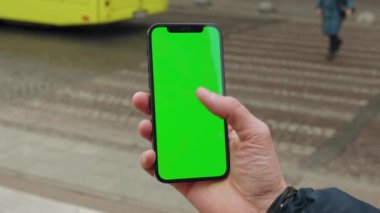 Arka plandaki bulanık yaya geçidinde yeşil ekranlı telefonu kullanan ve dinleyen bir adamın resmi. Özel uygulama tıklatma kaydırmasını göster. Açık havada. Yavaş çekim