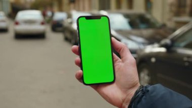 Elinde maket tutan genç adam, sokakta dikey yeşil ekranlı telefona dokunuyor ve araklıyor. Sosyal ağlar. Açık havada. Uygulama cep telefonu. Yavaş çekim