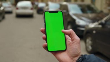Elinde manken tutan bir adamın sokakta dikey yeşil ekranlı telefona dokunurken ve araklarken görüntüsü. Arka plan arabası. Sosyal ağlar. Açık havada. Uygulama cep telefonu. Yavaş çekim