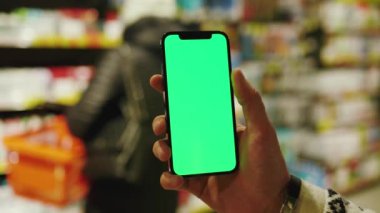 Yavaş çekimde çalışan adam, arka planda bulanık gece ışığı süpermarketinde dikey yeşil ekranlı cep telefonu kullanıyor. Model dinleme görüntülü kromakey telefon. Kapat.