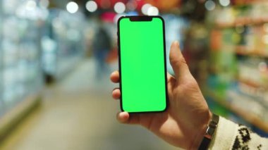 Kapat ellerini adamım, arka planda bulanık ürünler bulunan süpermarket raflarında dikey yeşil ekranlı cep telefonu kullan. Model dinleme görüntülü kromakey telefon. Yavaş çekim