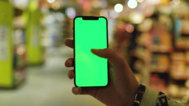 Akşamları hafif çalışan adam arka planda bulanık ürünler bulunan süpermarket raflarında dikey yeşil ekranlı cep telefonu kullanır. Model dinleme görüntülü kromakey telefon. Yavaş çekim