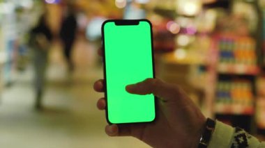 Eller, arka planda bulanık ürünler bulunan süpermarket raflarında dikey yeşil ekranlı cep telefonu kullanır. Model dinleme görüntülü kromakey telefon. Yavaş çekim