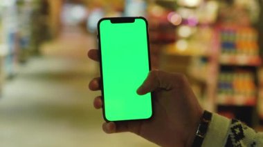 Kapat ellerini adamım, arka planda bulanık ürünler bulunan süpermarket raflarında dikey yeşil ekranlı cep telefonu kullan. Model dinleme görüntülü kromakey telefon. Yavaş çekim