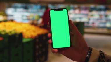 Elle çekilen adam arka planda bulanık ürünler içeren süpermarket raflarında dikey yeşil ekranlı cep telefonu kullanıyor. Model dinleme görüntülü kromakey telefon. Yavaş çekim